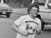 42nd Terry Fox Walk on September 18th a black and white photo of a white man with curly hair, a tshirt and shorts running along a paved road. His right leg is a prosletic.