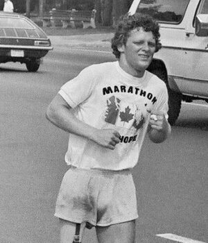 a black and white photo of a white man with curly hair, a tshirt and shorts running along a paved road. His right leg is a prosletic.